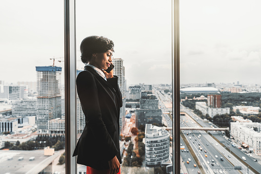 A serious African-American businesswoman is talking on the phone while standing near a window, on a top floor of a luxury business high-rise, with cityscape, construction site, and highroad outside