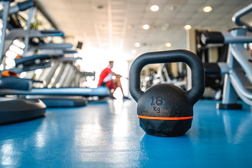 Low angle view of kettlebell sitting on gym floor and young male relaxing on treadmill after workout in background.