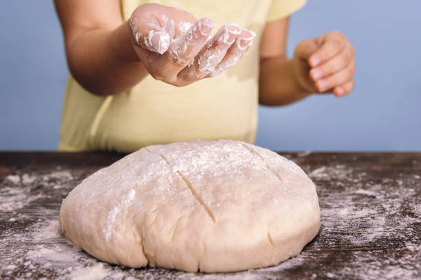 hands of an unrecognizable child putting flour on a rustic bread dough on a wooden table, concept of healthy food at home
