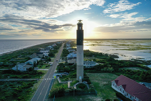 The sun is setting behind the Oak Island NC lighthouse. Surrounded by the ocean and waterway on a partly cloudy day.