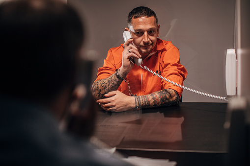 Two men, male prisoner talking over the phone with his lawyer in prison visit room.