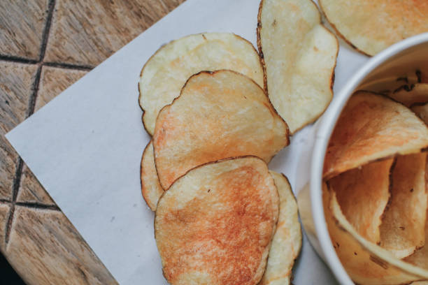 istock Fresh homemade deep fried crispy potato chips in white box on a wooden background, top view. Salty crisps scattered on a table for a tasty snack break. 1250153381