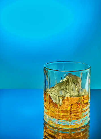 Crystal round glass with ice of scotch whiskey or brandy on a blue gradient background with reflection. Copy space
