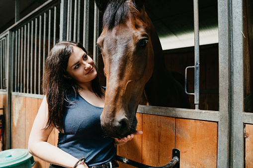 Young adult woman next to her horse in a horse riding school. The horse is peeking through his window.