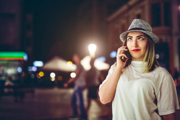 Young pretty Caucasian hipster woman with hat making a silly face while talking on phone downtown at night.