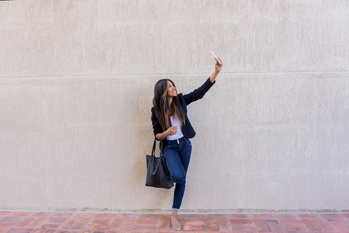 Young entrepreneur looking very happy taking a selfie against a wall outdoors using her cell phone - lifestyle concepts