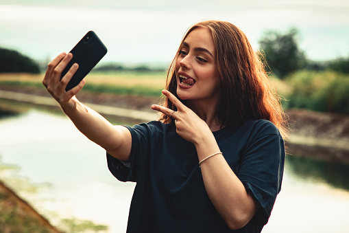 Joking around in front of the camera. Young woman standing outdoors vlogging and taking selfies with her mobile phone, smiling into the smart phone camera, joking around, showing peace hand sign. Outdoor Youth Social Media Lifestyle.