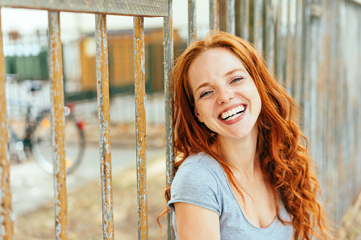 Pretty vivacious young redhead woman grinning at camera as she stands alongside a metal railing in town