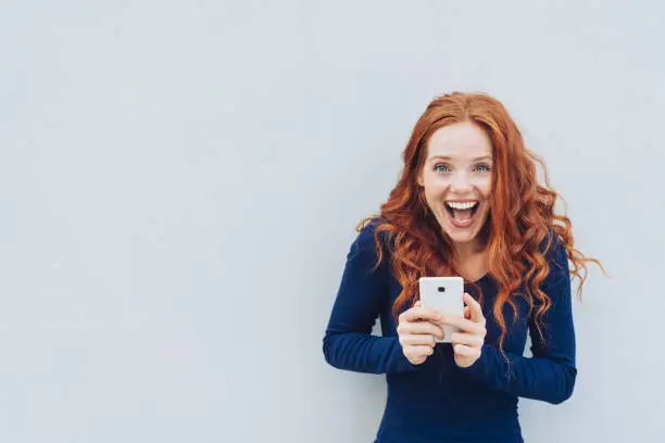 Vivacious young woman laughing at a good joke as she stands against a white wall with copy space holding a mobile