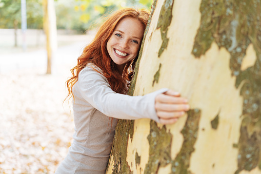 pretty redhead woman hugging a large tree looking around the trunk at the camera with a lovely friendly smile