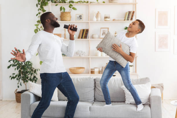 Cheerful black dad and preteen son having fun together in living room Home Entertainment. Cheerful black dad and preteen son having fun together in living room, singing and dancing, boy using pillow as guitar father and son guitar stock pictures, royalty-free photos & images