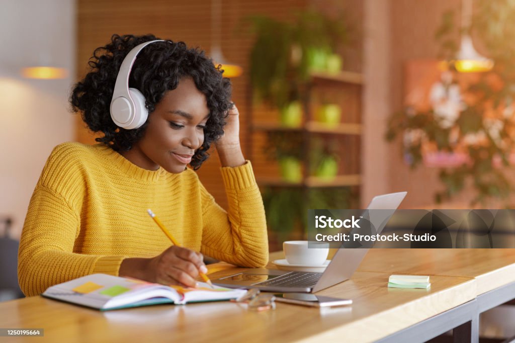 Smiling black girl with headset studying online, using laptop Smiling black girl with wireless headset studying online, using laptop at cafe, taking notes, copy space Learning Stock Photo