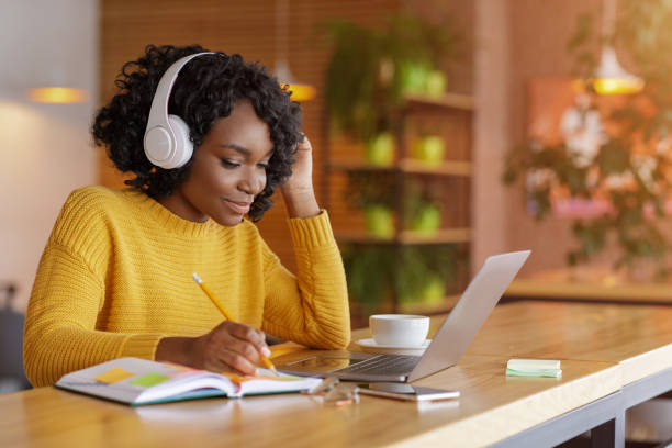 Smiling black girl with headset studying online, using laptop Smiling black girl with wireless headset studying online, using laptop at cafe, taking notes, copy space listening stock pictures, royalty-free photos & images