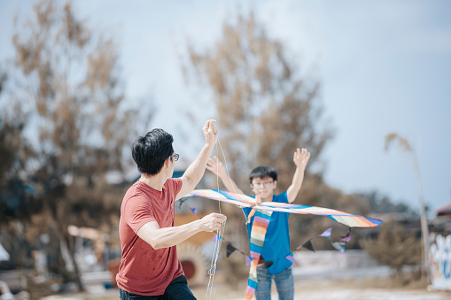 an asian chinese father playing and flying kite with his son at the beach