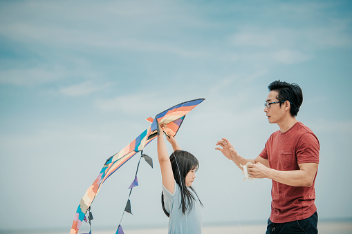 an asian chinese father playing and flying kite with his daughter at the beach