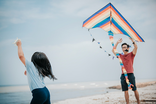 an asian chinese father playing and flying kite with his daughter at the beach