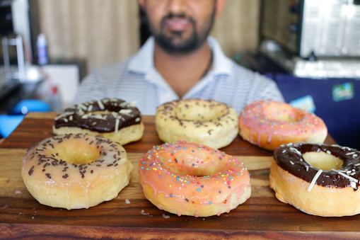 Stock photo showing unrecognised man holding home made fondant iced ring doughnuts in a row topped with sprinkles on wooden chopping board. The chopping board is being held by an unrecognisable person.