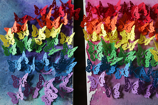 Stock photo of close-up of gay themed homemade crafting art work, using rainbow butterfly punch with cut out card and paper butterflies, embossed and glued on abstract watercolour painting that has been painted on a stretched canvas. This original craft art work of rainbow coloured made of multicoloured cut out butterfly shapes (Monarch paper butterflies - purple, blue, green, orange, yellow and red colours) is like the rainbow LGBTQI gay flag, with the colourful arts and crafts project displayed on wall as background wallpaper.
