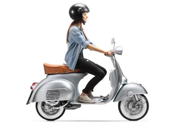Photo of Full length profile shot of a young woman with a helmet riding a vintage scooter