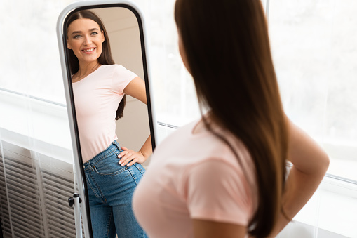 Perfect Weight. Happy Skinny Girl Looking At Reflection In Mirror Standing At Home. Successful Slimming Concept. Selective Focus