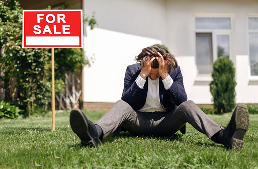Depressed real estate agent sitting on grass in front of house for sale, outside