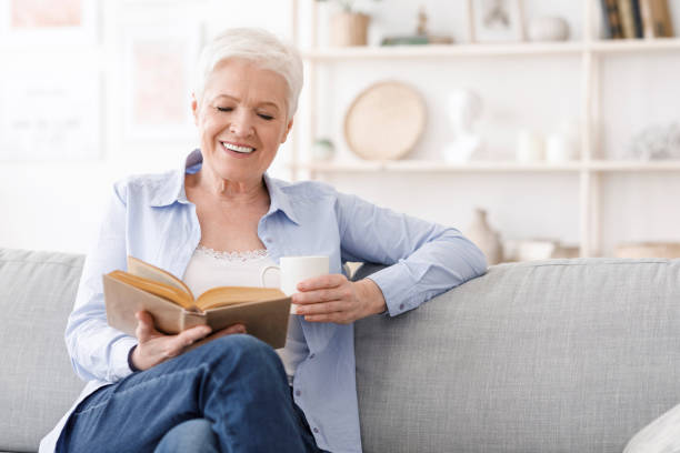 istock Retirement time. Elderly lady sitting on couch with book and drinking coffee 1250218442