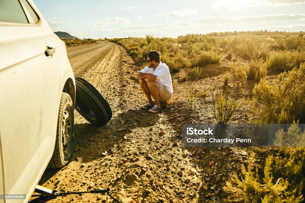Nothing to do but sit and wait for some roadside assistance Shot of a young man sitting on the roadside next to his car with a flat tyre in a rural area Stranded Stock Photo