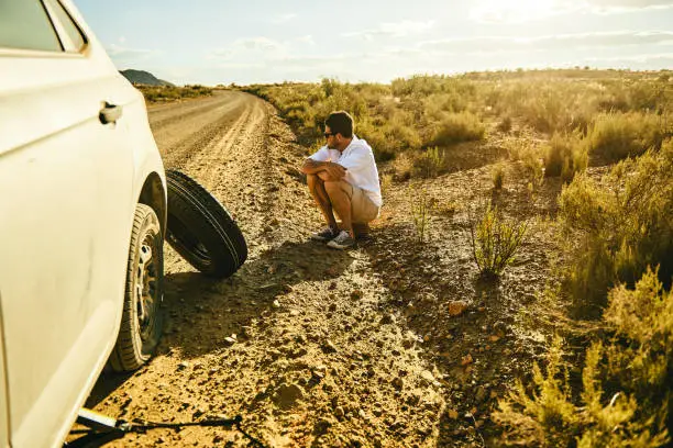 Shot of a young man sitting on the roadside next to his car with a flat tyre in a rural area