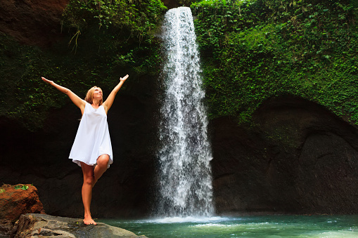 Young woman travel in Bali rainforest. Happy girl enjoy jungle nature. Stand in natural pool under waterfall, see on falling water. Walking day tour, hiking activity adventure on family summer holiday