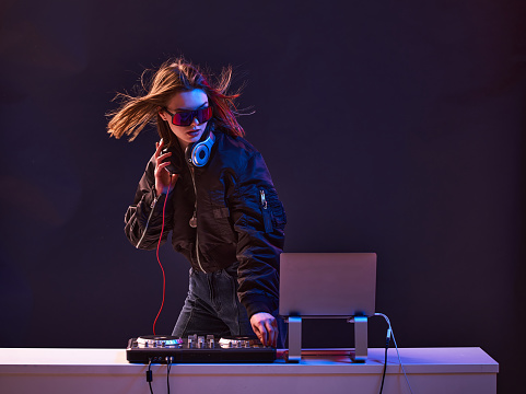 Stylish DJ girl with headphones and glasses on the bomber jacket mixes the music at the party and neon lights. High quality photo