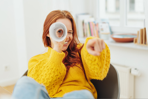 Playful young woman playing I Spy with the camera peering through rolled paper and pointing with focus to her face