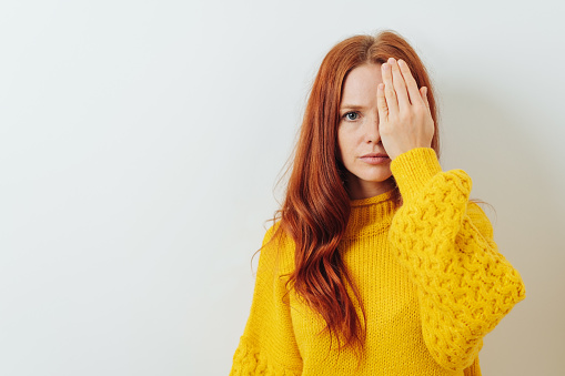 Young redhead woman covering one eye with her hand as she stares solemnly at the camera