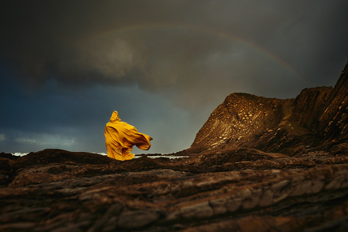 Low angle view of woman standing on stone wearing yellow cape, rainbow in the background