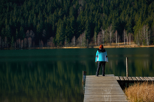 Rear view of redhead woman wearing blue coat looking at the view at the lake's forest; standing on the wooden pontoon