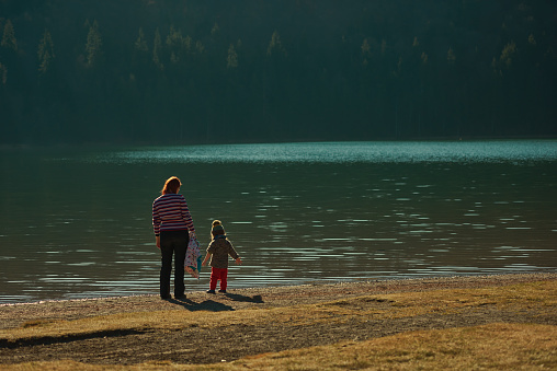 Rear view of redhead mother and baby daughter at the lake, investigating, looking down, body of water in the background