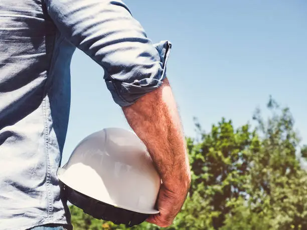Handsome engineer, holding white hardhat in the park against the backdrop of green trees and the setting sun, looking into the distance. Close-up. Concept of labor and employment