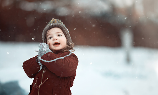 Little cute girl in white hat,scarf snood is lying,playing in snow. Kid walking in beautiful frozen forest, park among fir snowy trees. Winter vacation entertainment in village country house backyard.