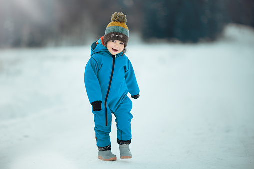 Cute child wearing a warm hat catching snowflakes with his tongue. Funny little boy in blue winter clothes walks during a snowfall. Outdoors winter activities for kids.