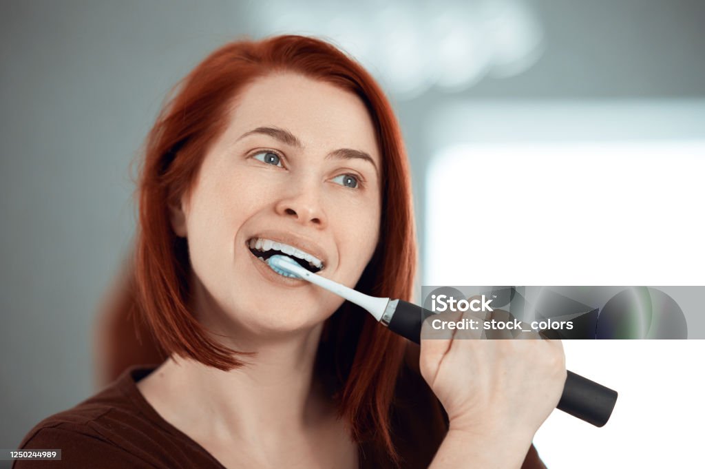 Loving the morning routine Portrait of redhead brushing her teeth with electric toothbrush, looking away and feeling fresh and clean Electric Toothbrush Stock Photo