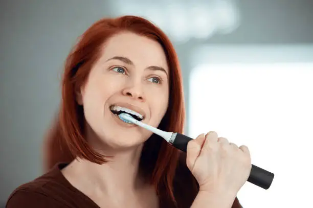 Portrait of redhead brushing her teeth with electric toothbrush, looking away and feeling fresh and clean