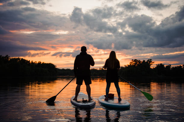 rear view on couple of people on sup boards on the river at sunset rear view on couple of people standing on sup boards with oars in their hands on the river at sunset paddleboard photos stock pictures, royalty-free photos & images