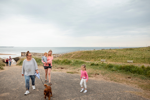A same sex couple walking away from the beach with their daughter and son, one woman is carrying their son as the other woman is holding their pet dog on a leash.