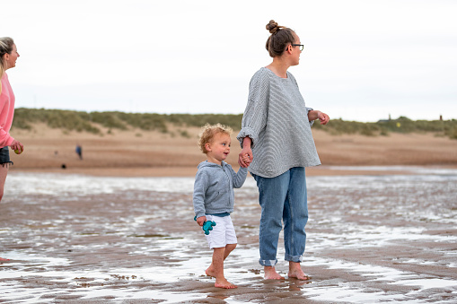 A woman holding hands with her son while they stand barefoot on the beach. Her fiancé is also standing behind them, slightly out of shot. They are all looking to their side.