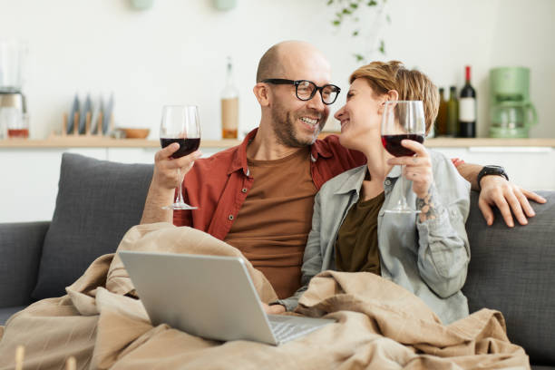 Couple spending time at home Happy mature couple sitting on sofa with glasses of red wine using laptop computer and laughing couple drinking stock pictures, royalty-free photos & images