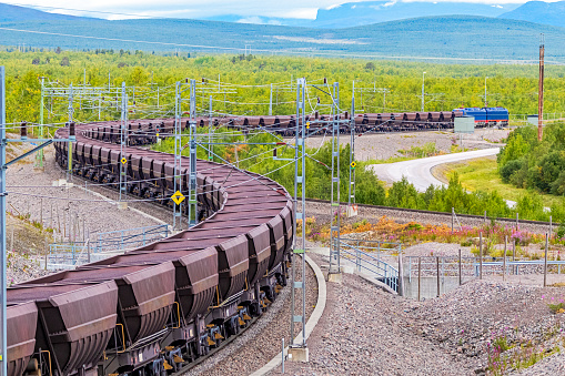 Loaded iron ore train snaking around curves through maze of electric poles and wires in Arctic Circle extreme terrain hauled by two electric locomotives: ID & logos removed, colour altered.