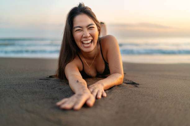 junge frau im bikini liegt auf ihrer vorderseite am strand und lacht glücklich - healthy lifestyle women beach looking at camera stock-fotos und bilder