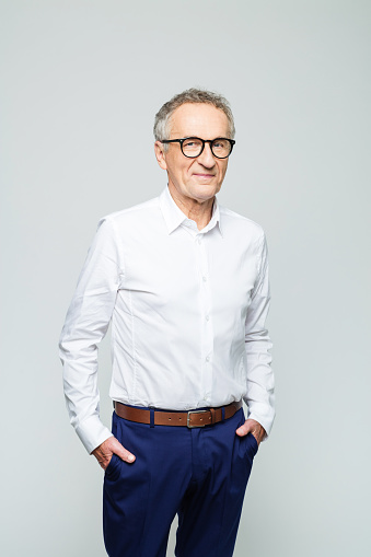 Portrait of elderly elegant man wearing white shirt and glasses smiling at camera. Confident senior businessman, studio shot against grey background.
