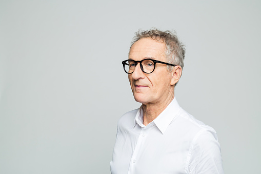 Portrait of elderly elegant man wearing white shirt and glasses looking away. Confident senior businessman, studio shot against grey background.