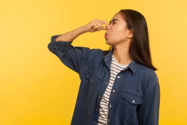 Awful smell! Portrait of girl in denim shirt grimacing in disgust and holding breath, grabbing nose with fingers, avoiding stinky odor, fart gases. indoor studio shot isolated on yellow background