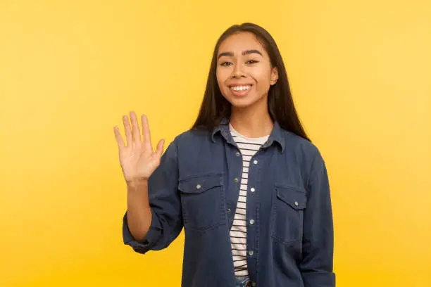 Photo of Hi, welcome! Portrait of friendly happy girl in denim shirt waving hand gesturing hello or goodbye, greeting with smile